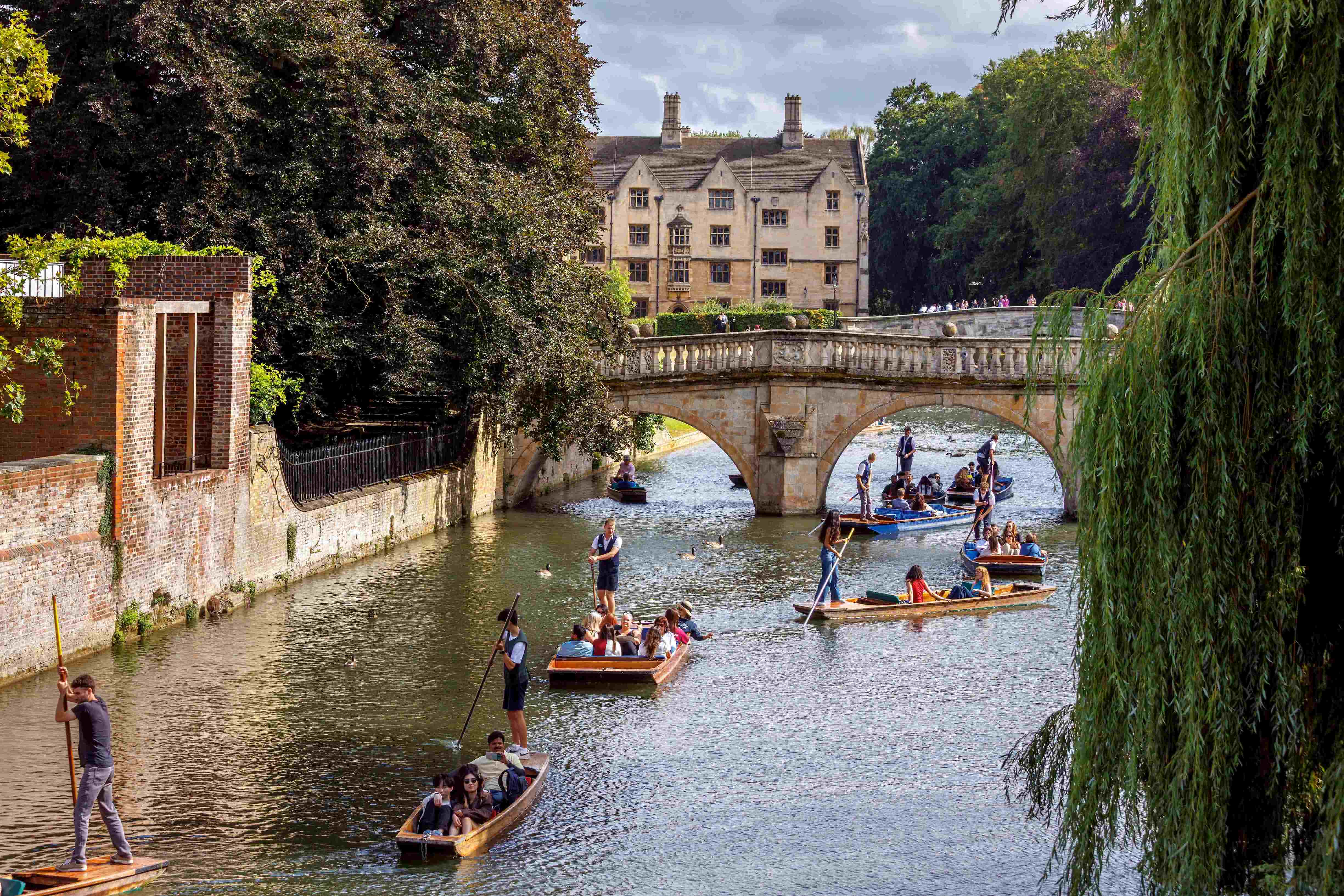 Punters guide boats along the River Cam beneath a stone bridge near Cambridge college buildings
