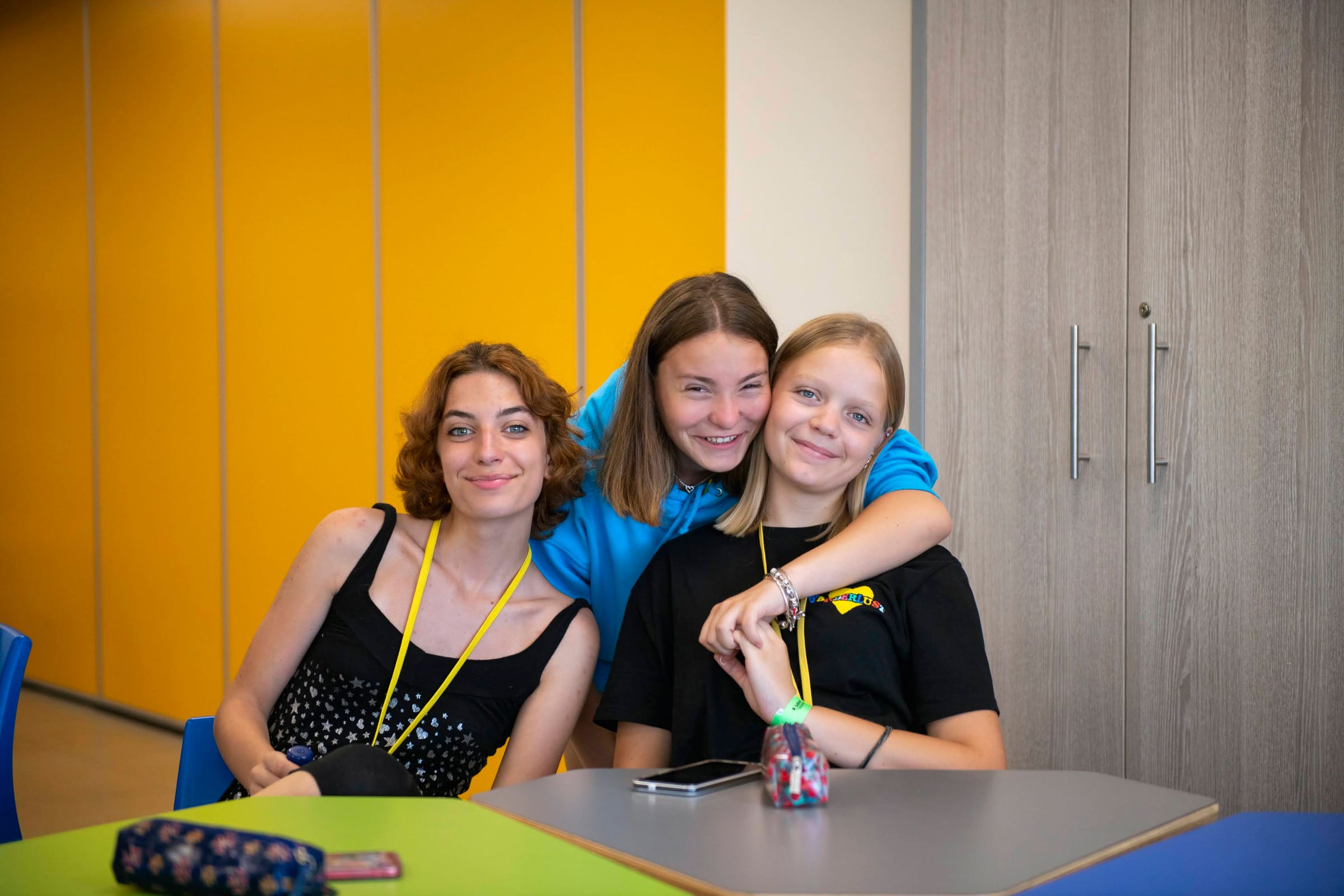 Three teenage girls with lanyards smiling together in a bright classroom during a Malta junior language course